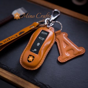 May include: A brown leather key fob with a silver key ring and a leather charm in the shape of the letter A. The key fob has a small, orange leather patch with the letter M on it. The key fob is designed to fit a car key.