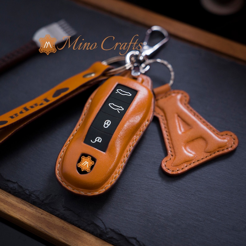 May include: A brown leather key fob with a silver key ring and a leather charm in the shape of the letter A. The key fob has a small, orange leather patch with the letter M on it. The key fob is designed to fit a car key.