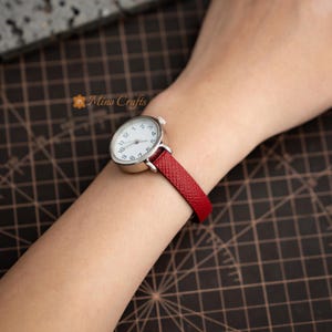 Peut inclure: Une petite montre-bracelet ronde argentée avec un cadran blanc et des chiffres noirs. La montre a un bracelet en cuir rouge. La montre est sur un poignet. Le logo Mino Crafts est dans le coin supérieur gauche.