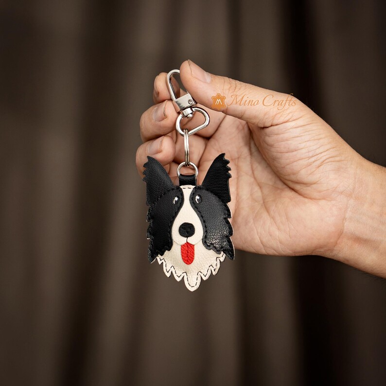 May include: A black, white, and red leather keychain in the shape of a Border Collie dog. The keychain features a silver-colored clasp and a key ring. The dog's face has a white muzzle, black ears, and a red tongue.