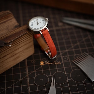 May include: A small, round wristwatch with a white face and silver casing. The watch has a vibrant orange leather band. The watch is displayed on a wooden surface, with crafting tools in the background.