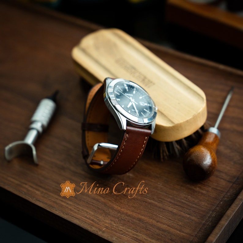 May include: A brown leather watch strap with a silver watch face and hands. The watch is on a wooden surface with a brush and a leather tool.