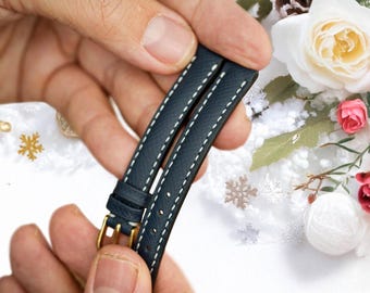 May include: A dark blue leather watch strap with white stitching. The strap is folded in half and held by a hand.