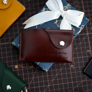 May include: A brown leather wallet with a silver button and an engraved quote: "You may delay, but time will not".