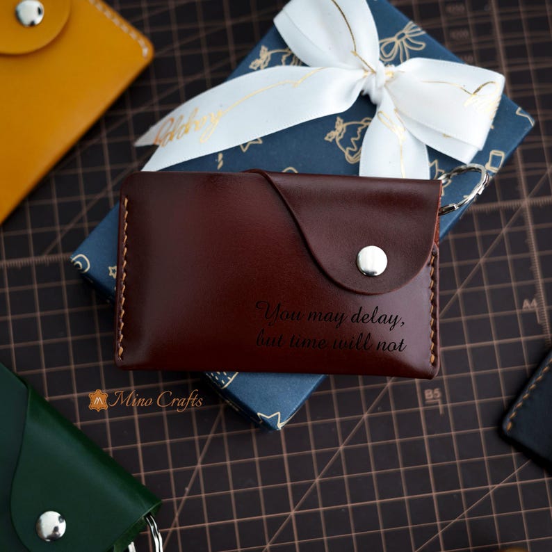 May include: A brown leather wallet with a silver button and an engraved quote: "You may delay, but time will not".