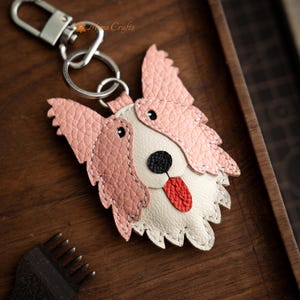 May include: A handcrafted leather dog keychain with a pink and white design. The dog's face features a black nose, red tongue, and black eyes. The keychain includes a silver clasp and ring.