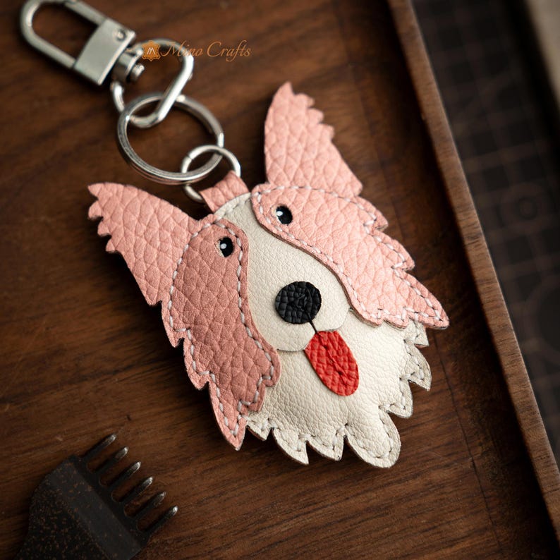 May include: A handcrafted leather dog keychain with a pink and white design. The dog's face features a black nose, red tongue, and black eyes. The keychain includes a silver clasp and ring.