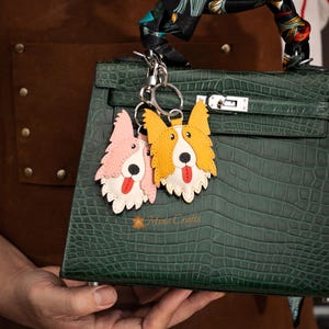 May include: Two dog-shaped keychains, one pink and one yellow, with white and red accents, hang from a silver keyring. The keychains are attached to a dark green textured handbag. The text "Mina Crafts" is visible.
