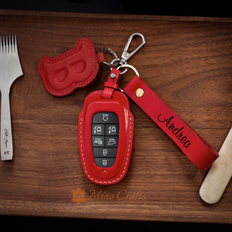May include: Red leather key fob with a red leather strap and the name "Andrea" engraved on it. The key fob has a red leather loop and a silver metal key ring.