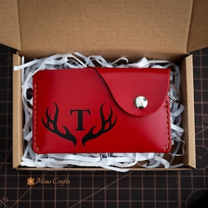 May include: A red leather key chain with a snap closure and a black antler design with the letter "T" in the center. The key chain is in a white shredded paper-filled box.