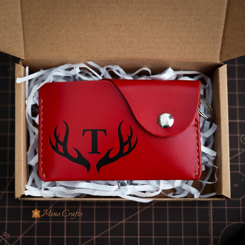 May include: A red leather key chain with a snap closure and a black antler design with the letter "T" in the center. The key chain is in a white shredded paper-filled box.