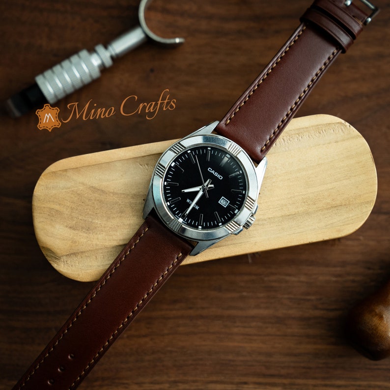 May include: A brown leather watch strap with a silver watch face that reads "CASIO" and has a black background. The watch is on a wooden surface.