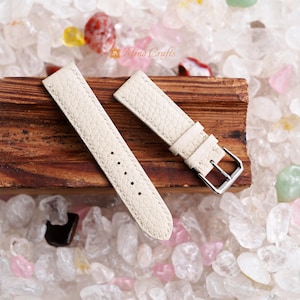 Könnte beinhalten: Zwei cremefarbene Lederarmbänder mit einer silbernen Schließe. Die Armbänder liegen auf einer Holzoberfläche mit weißen Steinen drumherum.
