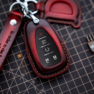 Op de afbeelding: Een rode leren sleutelhanger met een zwarte sleutel erin. De sleutelhanger heeft een gestikte rand en een kleine metalen lus om aan een sleutelring te bevestigen. De sleutel heeft vier knoppen met zilveren pictogrammen.