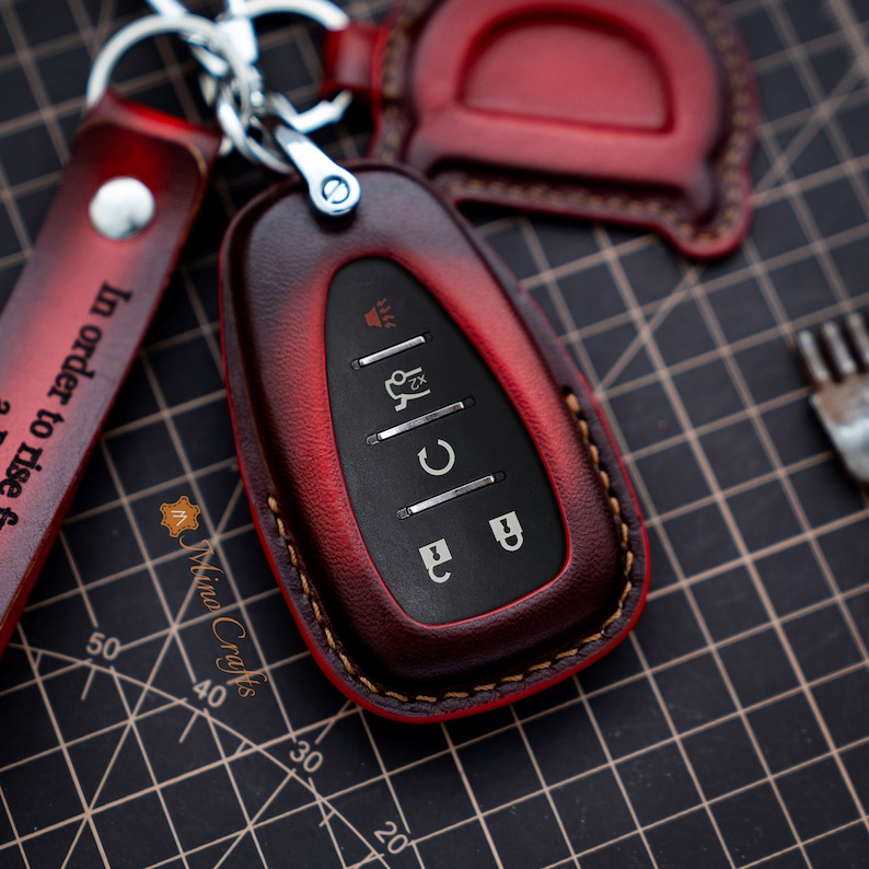 Op de afbeelding: Een rode leren sleutelhanger met een zwarte sleutel erin. De sleutelhanger heeft een gestikte rand en een kleine metalen lus om aan een sleutelring te bevestigen. De sleutel heeft vier knoppen met zilveren pictogrammen.