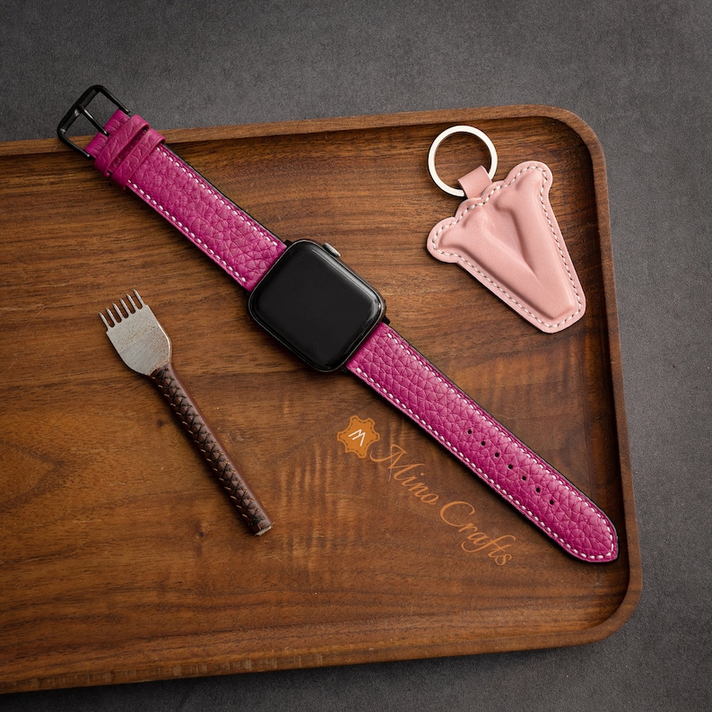 May include: A pink leather watch strap with white stitching and a black buckle. The strap is on a wooden tray with a leather key chain with a pink letter V.