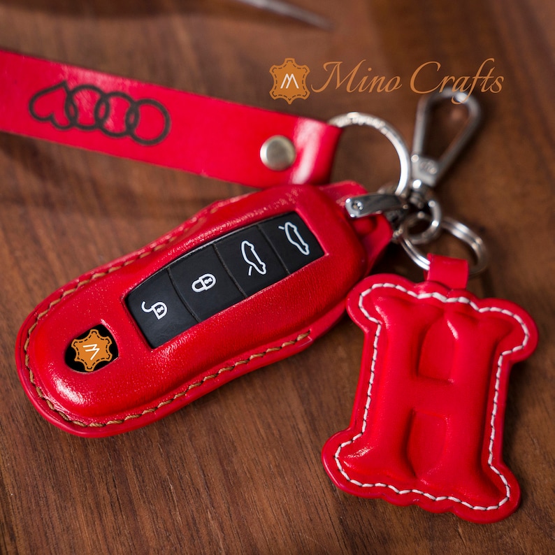 May include: Red leather key fob with the Audi logo and a red leather keychain with the letter H. The keychain is stitched with white thread.