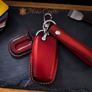 May include: Red leather key fob with a stitched edge and a matching leather strap with a silver metal clasp. The key fob has a small leather loop on the side. The text "Mino Crafts" is visible in the background.