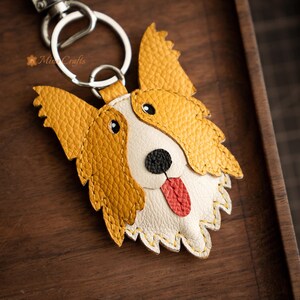 May include: A handcrafted leather keychain in the shape of a dog's face. The dog has yellow and white fur, black eyes, a black nose, and a red tongue. The keychain includes a silver-colored metal ring and clasp.