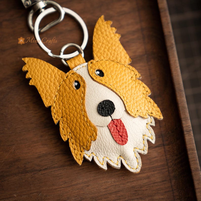 May include: A handcrafted leather keychain in the shape of a dog's face. The dog has yellow and white fur, black eyes, a black nose, and a red tongue. The keychain includes a silver-colored metal ring and clasp.