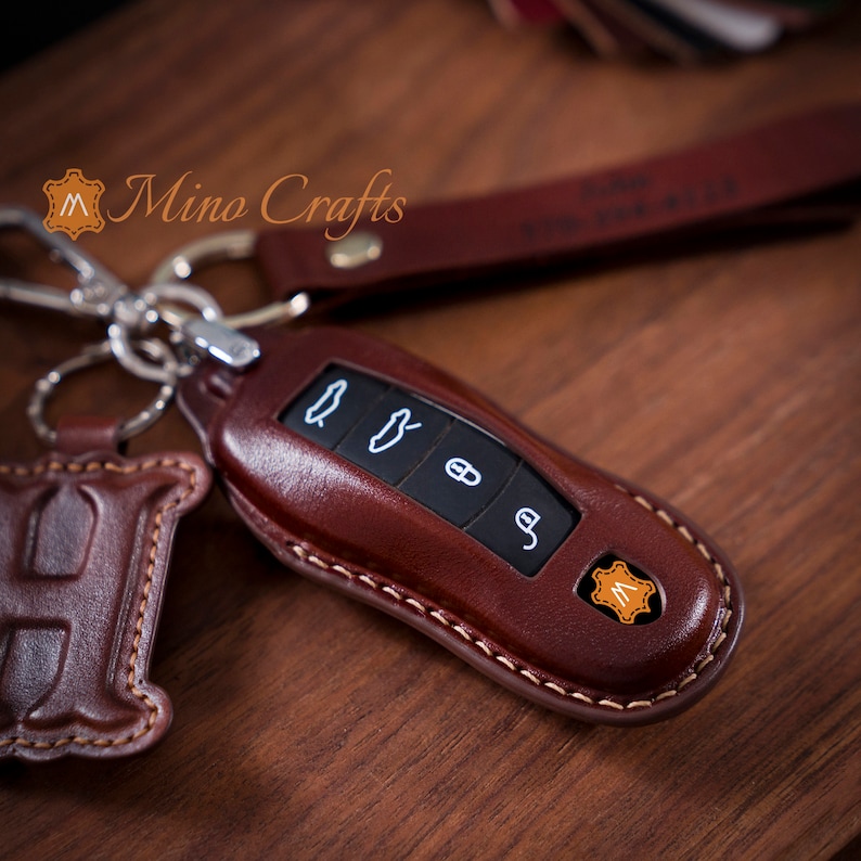 May include: A brown leather key fob with stitching around the edges. The fob has four buttons with symbols on them. The fob has a small leather tag with the letter "M" on it.