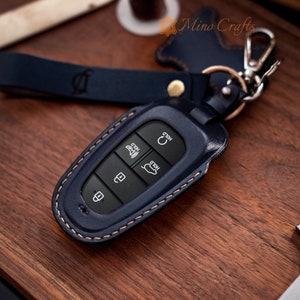 May include: A blue leather key fob with stitching detail and a silver key ring. The fob has four buttons labeled "Hold" and a single button with a car icon. The fob is on a wooden surface.