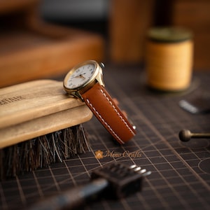 Peut inclure: Une montre dorée avec un cadran blanc et des chiffres romains. La montre est dotée d'un bracelet en cuir marron avec des coutures blanches. La montre est présentée sur une surface en bois, avec une brosse et d'autres outils en arrière-plan.