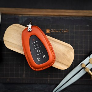 Op de afbeelding: Oranje leren sleutelhanger met stiksels rond de randen. De sleutelhanger heeft een zwarte sleutel met knoppen voor vergrendelen, ontgrendelen, kofferbak en paniek.