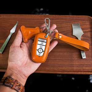 May include: A brown leather key fob with a silver car key inside. The key fob has a loop for attaching to a keychain. The key fob is on a wooden surface.