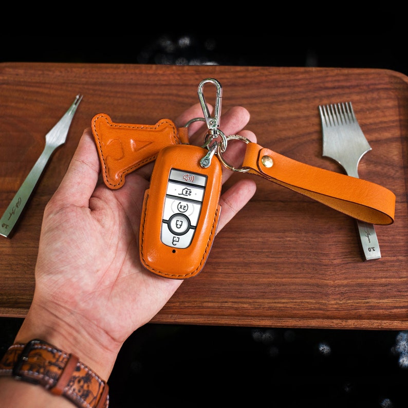 May include: A brown leather key fob with a silver car key inside. The key fob has a loop for attaching to a keychain. The key fob is on a wooden surface.
