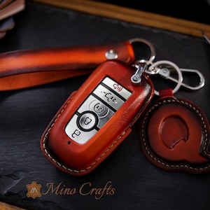 May include: A close-up of a red leather key fob with a silver key chain. The key fob has a silver button with a lock icon and a silver button with a car icon. The key fob is on a black surface.