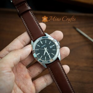 May include: A brown leather watch strap with white stitching, attached to a silver watch with a black face and silver hands. The watch face has the word "CASIO" printed on it.