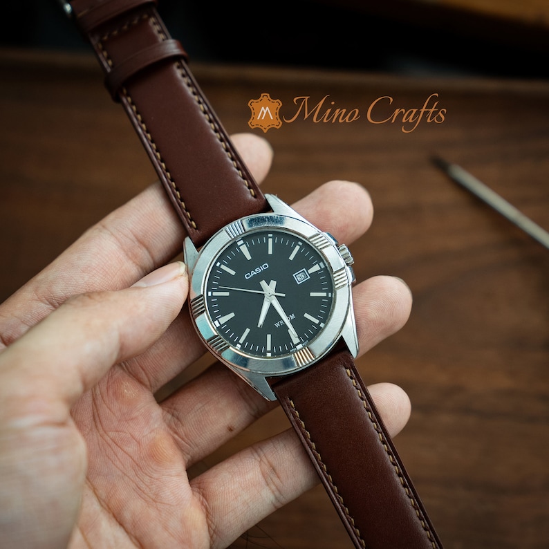 May include: A brown leather watch strap with white stitching, attached to a silver watch with a black face and silver hands. The watch face has the word "CASIO" printed on it.