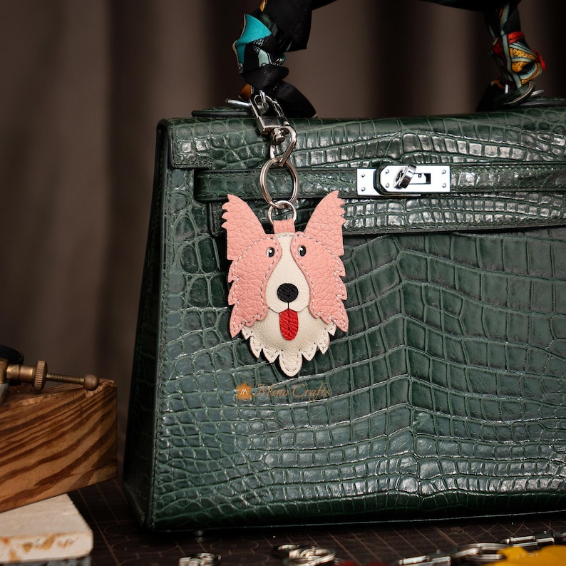 May include: A pink and white leather dog keychain with a red tongue, hanging from a silver keyring. The dog keychain is attached to a dark green leather handbag with a crocodile skin pattern.