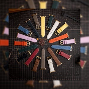 Pode incluir: Uma coleção de pulseiras de relógio de couro coloridas, em várias tonalidades, incluindo azul, laranja e verde, dispostas sobre uma superfície escura. Cada pulseira tem uma fivela dourada e é projetada para um smartwatch. As pulseiras têm diferentes texturas.