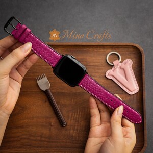 May include: A pink leather watch strap with white stitching, attached to a black smartwatch. The strap is shown on a wooden surface with a small pink leather keyring in the background.