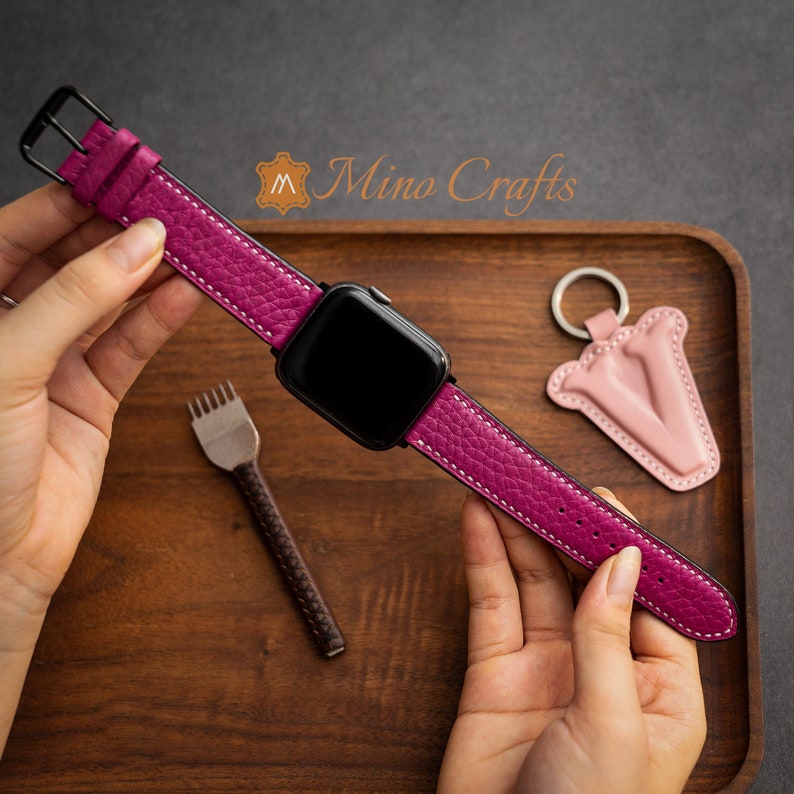 May include: A pink leather watch strap with white stitching, attached to a black smartwatch. The strap is shown on a wooden surface with a small pink leather keyring in the background.
