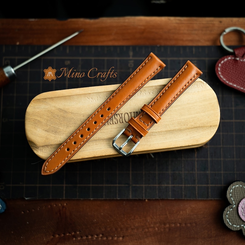 May include: Two brown leather watch straps with stitching detail. One strap has a silver buckle. The straps are on a wooden surface.