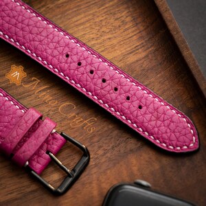 May include: A pink leather watch strap with white stitching. The strap has a black buckle and is shown on a wooden surface.
