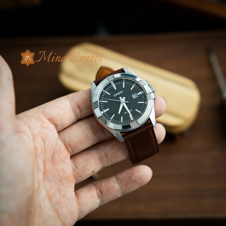 May include: A silver Casio watch with a black face and brown leather band. The watch has a date window and a silver bezel with markings.