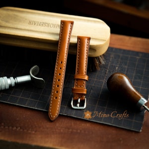 May include: Two brown leather watch straps with stitching detail. The straps are laid out on a black grid surface with a wooden brush and a leather tool.