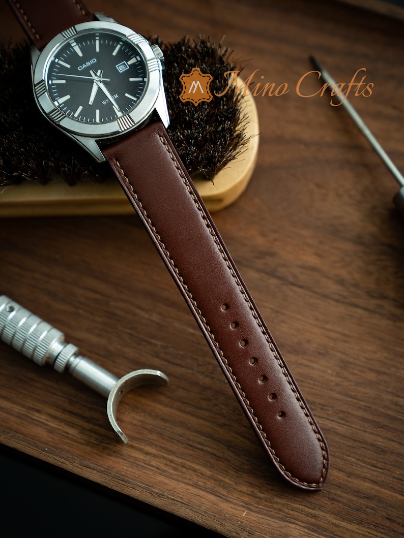 May include: A brown leather watch strap with a stitched edge and multiple holes for adjusting the fit. The strap is attached to a silver watch with a black face and the word "CASIO" printed on it.