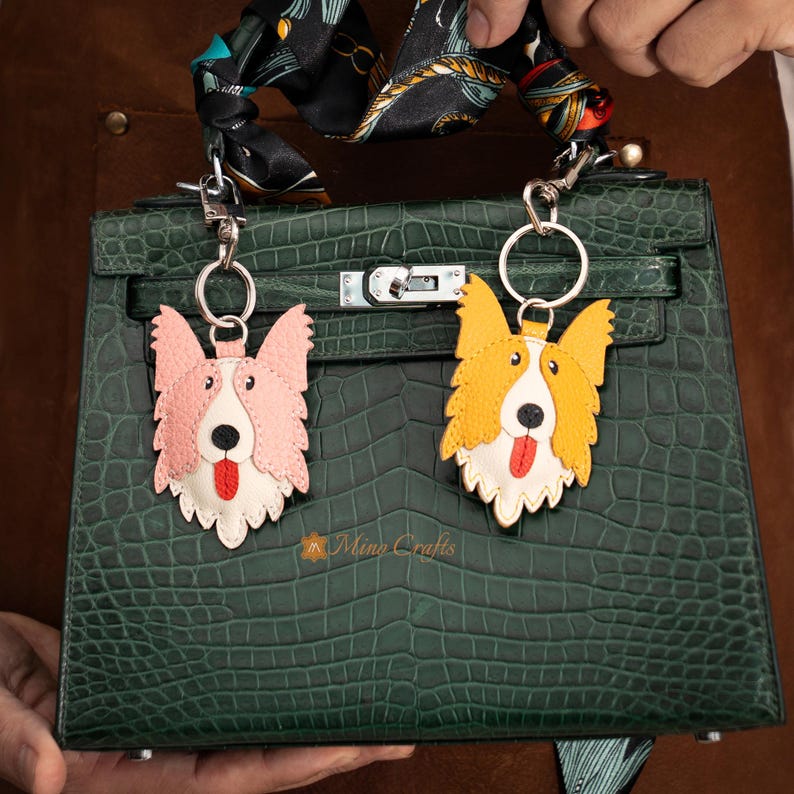 May include: Two leather dog keychains, one pink and one yellow, with white and red accents, hang from a green handbag. The keychains are shaped like corgis, with their tongues sticking out. The handbag has a crocodile skin texture.