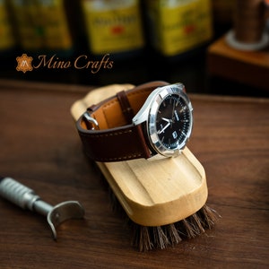 May include: A brown leather watch band with a silver watch face. The watch face has black hands and a black background with white markings. The watch is sitting on a wooden brush with brown bristles.