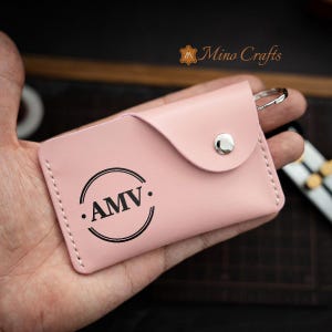 May include: A pink leather key holder with a silver snap closure and a metal ring. The key holder has a circular design with the initials "AMV" in black lettering.