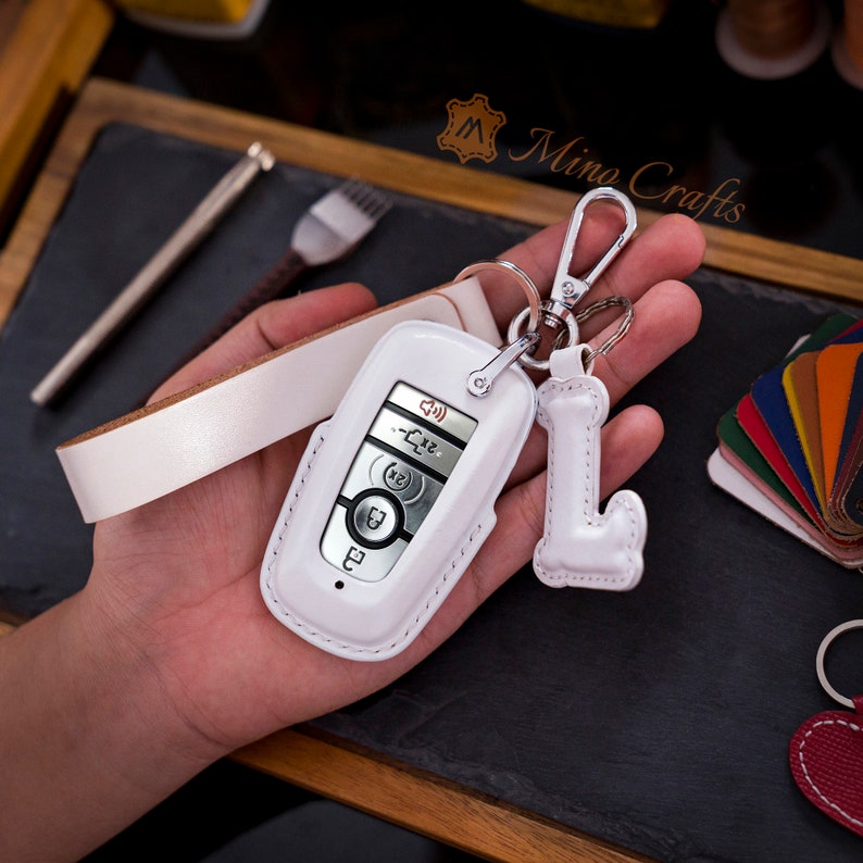 May include: White leather key fob with a white leather strap and a white leather letter L charm. The key fob has a silver key with buttons for lock, unlock, trunk, and panic.
