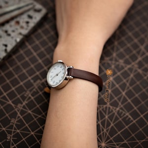 May include: A silver wristwatch with a white face and black numerals, paired with a dark brown leather band. The watch is displayed on a wrist, showcasing its classic design and elegant simplicity.