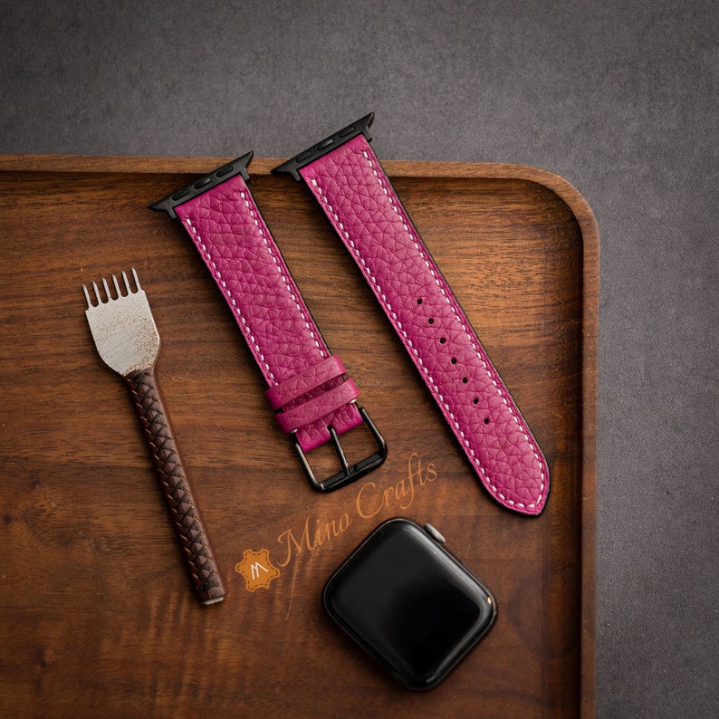 May include: Two pink leather watch straps with white stitching and black hardware on a wooden surface. The straps are for an Apple Watch.