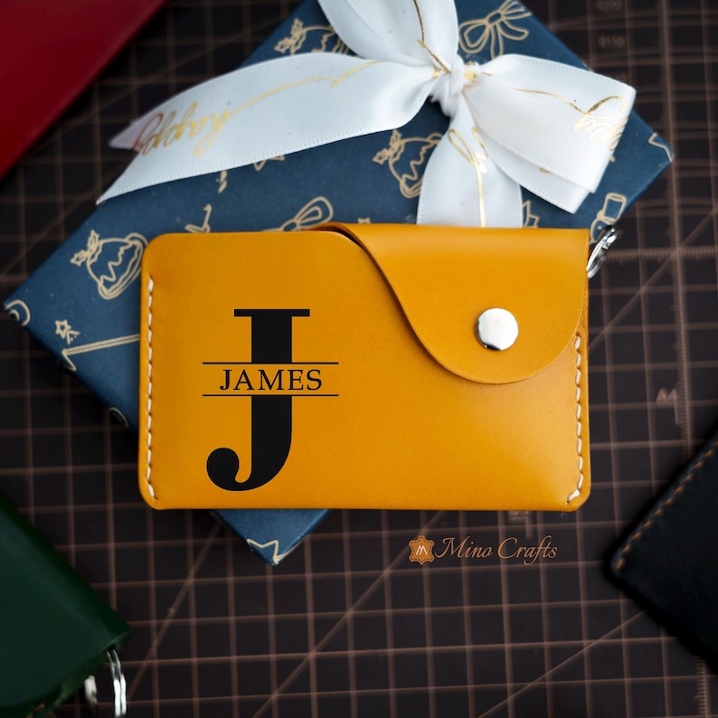 May include: A yellow leather card holder with a black letter "J" and the name "JAMES" engraved on the front. The card holder has a snap closure and a small loop for a keychain.
