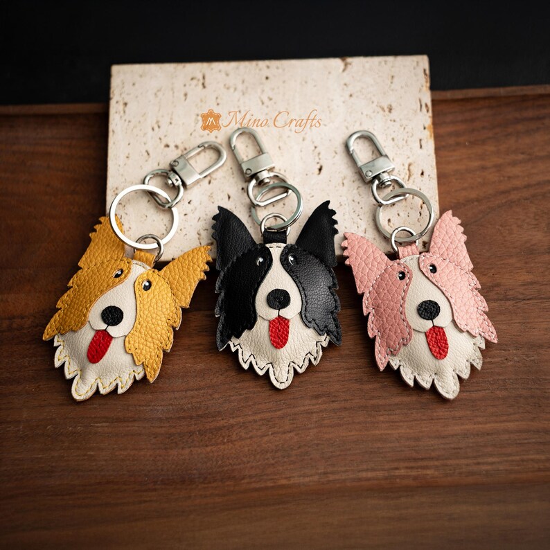 May include: Three leather dog keychains in yellow, black and white, and pink. Each keychain features a dog face with a red tongue, black eyes, and a silver keyring. The keychains are on a light-colored surface.